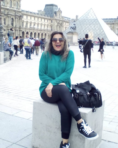 Foto de Sueli Furtado, sentada, rindo, em frente ao Museu do Louvre
